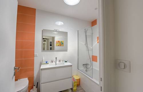 Salle de bains rénovée avec meuble vasque blanc, baignoire avec pare-douche et carrelage blanc et orange.