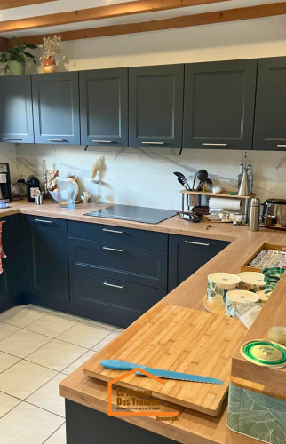 Vue d’ensemble d’une cuisine en L avec placards bleu nuit, crédence en marbre blanc et plafond à poutres apparentes.