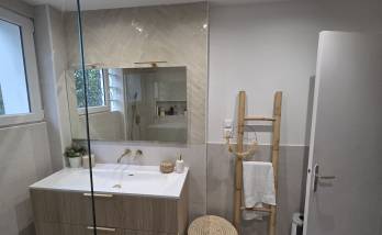 Salle de bains aux tons beiges avec meuble vasque effet bois, miroir mural, robinetterie dorée et carrelage mural contemporain.