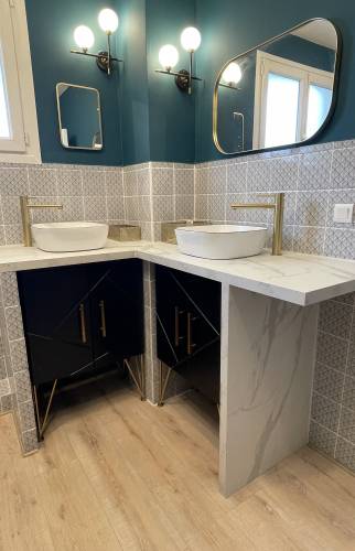 Zoom sur le coin double vasque d’une salle de bain rénovée à Carqueiranne, avec robinetterie dorée, faïence claire et meubles noirs sous plan en pierre blanche.