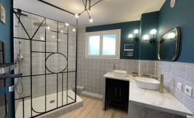 Vue d’ensemble d’une salle de bain rénovée à Carqueiranne, avec douche à l’italienne, verrière noire style atelier et éclairage moderne au plafond.