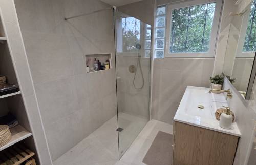 Salle de bains moderne avec douche à l’italienne vitrée, meuble vasque effet bois, carrelage beige et rangements intégrés.