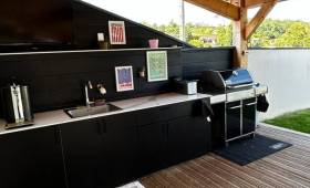 Cuisine d'été sur mesure finalisée à Coublevie avec rangements intégrés, évier, plan de travail et barbecue sous terrasse couverte.