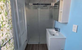Salle de bains moderne rénovée avec meuble vasque blanc, miroir rond et douche vitrée