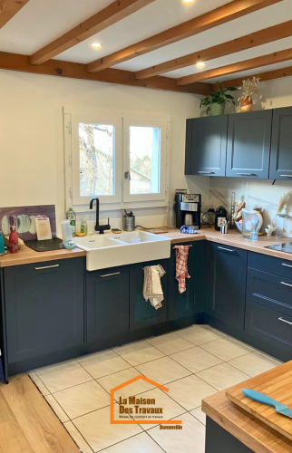 Évier blanc en céramique sous une fenêtre, meubles de cuisine bleu foncé, plan de travail en bois clair et électroménager moderne.