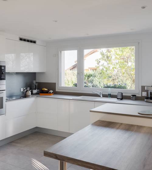 Cuisine rénovée avec meubles blancs brillants, plan de travail gris, évier sous la fenêtre et carrelage gris clair
