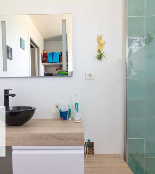 Vue d’ensemble lumineuse d’une salle de bain moderne comprenant une douche à l’italienne avec paroi vitrée et des carreaux vert émeraude. Le meuble suspendu en bois clair, surmonté d’une vasque noire design, apporte une touche d’élégance et de modernité.