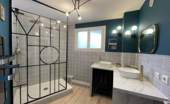 Vue d’ensemble d’une salle de bain rénovée à Carqueiranne, avec douche à l’italienne, verrière noire style atelier et éclairage moderne au plafond.