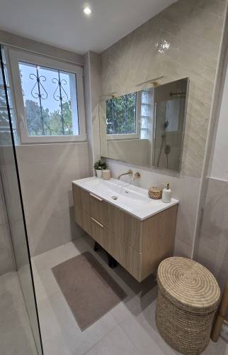 Meuble vasque suspendu effet bois avec plan blanc, grand miroir, carrelage beige et accessoires naturels dans une salle de bains moderne.