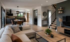Pièce de vie contemporaine avec cuisine noire et bois ouverte sur un salon lumineux, parquet en bois et escalier bois et métal.