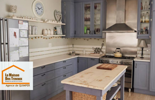 Cuisine rénovée au style campagne chic avec meubles bleu-gris, crédence claire et îlot central en bois patiné – un projet réalisé par La Maison Des Travaux Quimper.