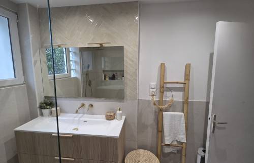 Salle de bains aux tons beiges avec meuble vasque effet bois, miroir mural, robinetterie dorée et carrelage mural contemporain.