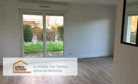 Un salon lumineux avec une grande baie vitrée donnant sur un espace extérieur verdoyant. Le parquet clair apporte une ambiance chaleureuse et moderne à la pièce, en harmonie avec les murs blancs qui accentuent la clarté de l'espace. 