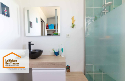 Focus sur le meuble de salle de bain suspendu, en bois clair, avec une vasque noire et un miroir rectangulaire. L’agencement soigné et minimaliste illustre parfaitement l’équilibre entre esthétique et fonctionnalité dans cette rénovation.