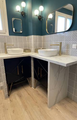 Zoom sur le coin double vasque d’une salle de bain rénovée à Carqueiranne, avec robinetterie dorée, faïence claire et meubles noirs sous plan en pierre blanche.
