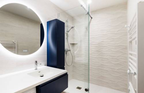 Salle de bains rénovée avec douche à l’italienne vitrée, meuble vasque bleu foncé, miroir rond rétroéclairé et carrelage mural texturé.