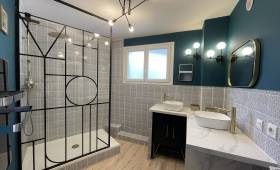 Vue d’ensemble d’une salle de bain rénovée à Carqueiranne, avec douche à l’italienne, verrière noire style atelier et éclairage moderne au plafond.