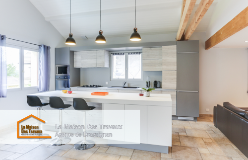 Une cuisine moderne avec un îlot central spacieux et élégant, idéal pour les repas en famille. Les suspensions noires apportent une touche de design, contrastant joliment avec les meubles en bois clair et gris.