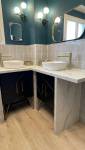 Zoom sur le coin double vasque d’une salle de bain rénovée à Carqueiranne, avec robinetterie dorée, faïence claire et meubles noirs sous plan en pierre blanche.