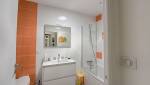 Salle de bains rénovée avec meuble vasque blanc, baignoire avec pare-douche et carrelage blanc et orange.